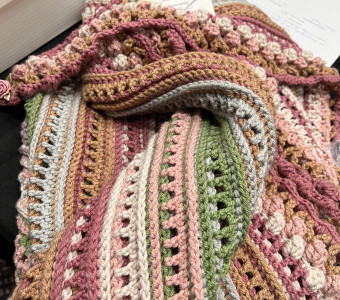 A handmade shawl or throw with stripes of earthy colors including pinks, greens, grays, and tans.