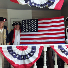Three men dressed in Revolutionary War uniforms stand on a porch dressed in red white and blue buntings while two of them display an American flag.
