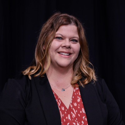 Image of Jamie Buster, wearing a red printed blouse and a black jacket against a black background.