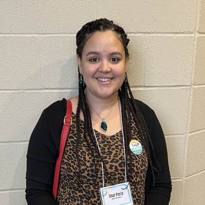 Image of Shai Perry wearing a cheetah print shirt, black cardigan, and nametag on a lanyard.