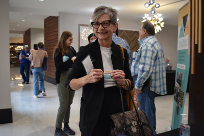 A woman dressed in black and white with black glasses, gray hair, and a bag smiles as she holds a High 5 1 Up Club pin.