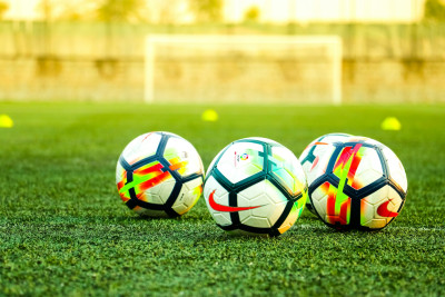 Image of a soccer pitch with goal in the background and four Nike soccer balls that are black and white with neon red and neon green highlights in the foreground.  Photo by Vikram TKV on Unsplash. 