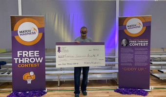 KMA Board Member Reese Barrick stands in a gym between Match Madness signs and holds a large check to KMA for $60 he won via a free throw contest.