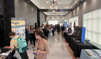 A long hotel hallway is filled with people and exhibit tables on both sides.