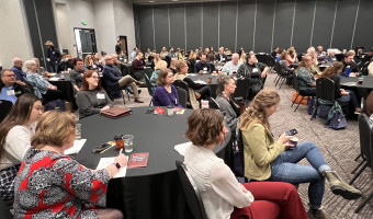 People sit in a large ballroom at round tables with black tablecloths.  They are looking to the right of the photo and listening to a speaker who is not pictured.