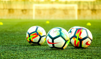 Image of a soccer pitch with goal in the background and four Nike soccer balls that are black and white with neon red and neon green highlights in the foreground.  Photo by Vikram TKV on Unsplash. 